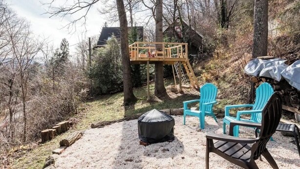 Una terraza de madera con una escalera, un hogar cubierto y sillas turquesas en un área boscosa.