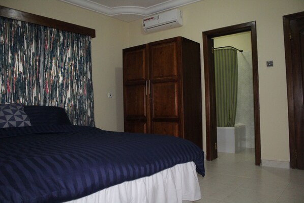 A bedroom with a bed, a wardrobe, a bathroom visible through an open door, and an air conditioning unit on the wall. - Evergreen Apartments (Adenta)