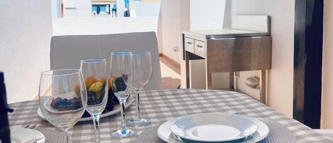 Mesa dispuesta para comer, con platos blancos, copas de vino y una botella, con vista a un edificio de paredes blancas y cielo azul.