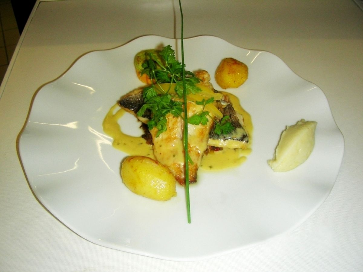 Un plato blanco con un filete de pescado, patatas y una guarnición sobre un mantel blanco.