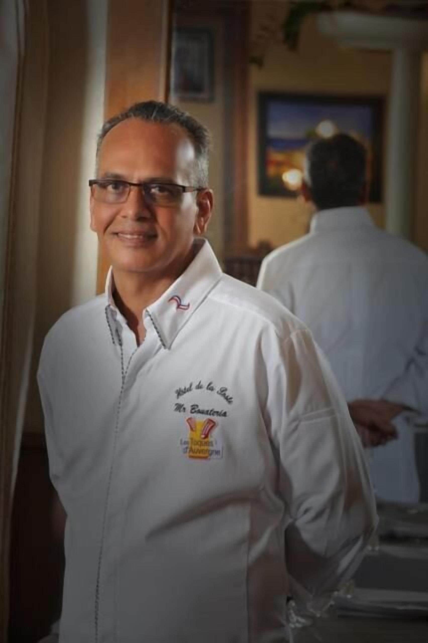 Una persona con camisa blanca que lleva un logo y texto, de pie en una habitación con un cuadro en la pared.