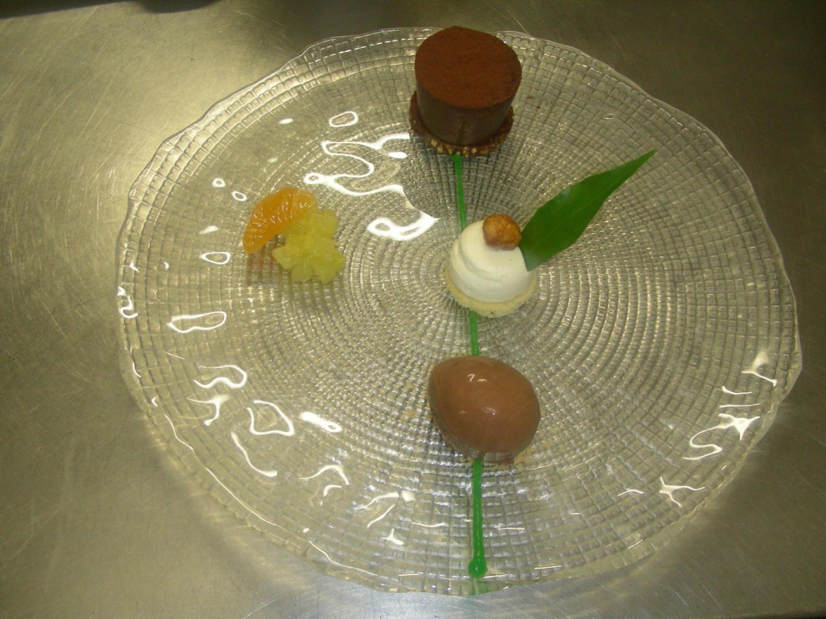 A dessert plate with four items: a chocolate sphere, a white sphere with a green leaf, a yellow fruit, and a brown sphere.