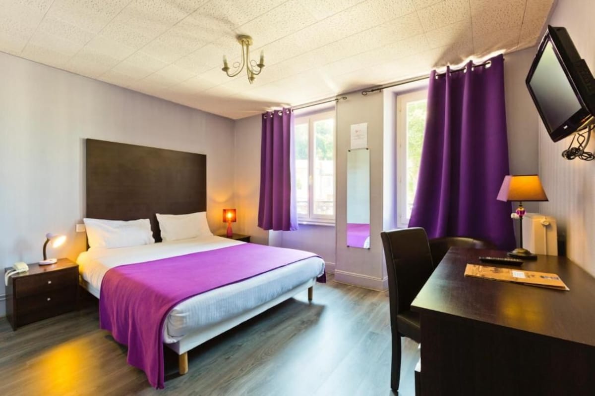 A hotel room with a bed, a desk, a chair, a TV, and a window with purple curtains.