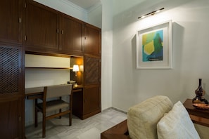 A hotel room with a wooden desk, a chair, a lamp, a framed abstract painting, and a sofa.