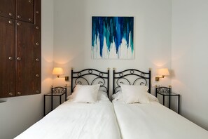 Een tweepersoonsbed met een witte deken, twee nachtkastjes met lampen, een wanddecoratie in abstractie en een houten garderobe.