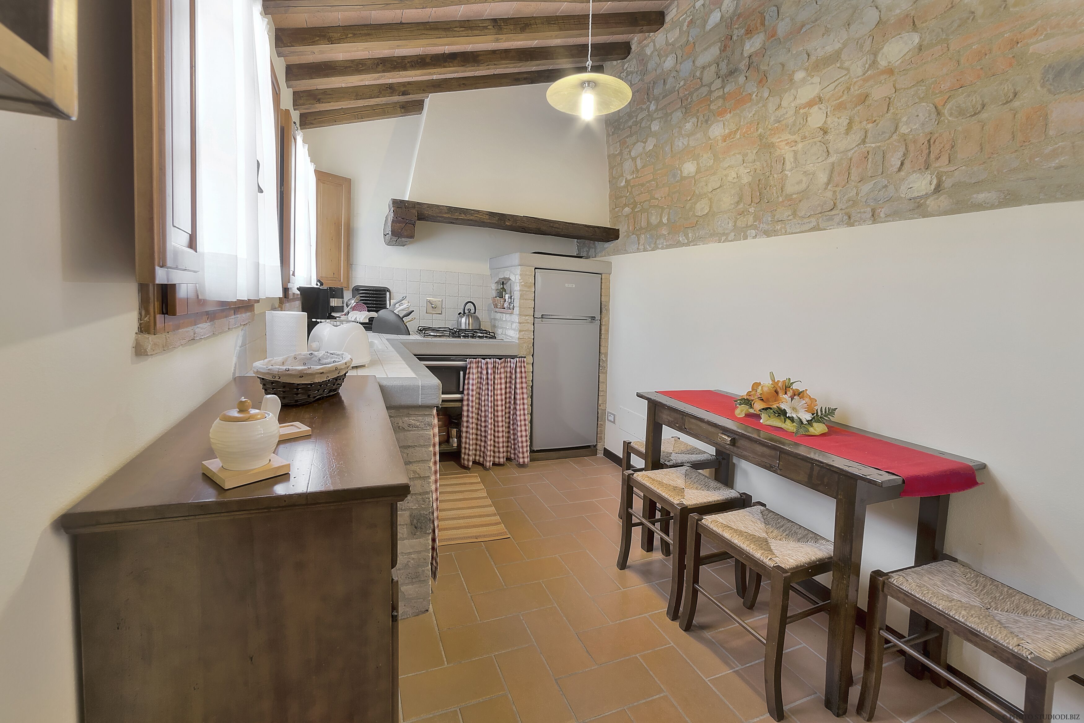 A kitchen with a stone wall, wooden beams, a red tablecloth, and a basket on the counter.