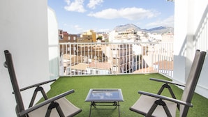Un balcon avec deux chaises et une petite table, d’où l’on peut admirer un paysage urbain avec des bâtiments et une montagne.