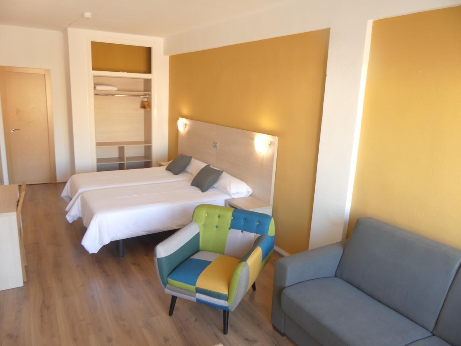 Camera d'albergo con due letti, una poltrona colorata, un divano grigio e pavimento in legno.