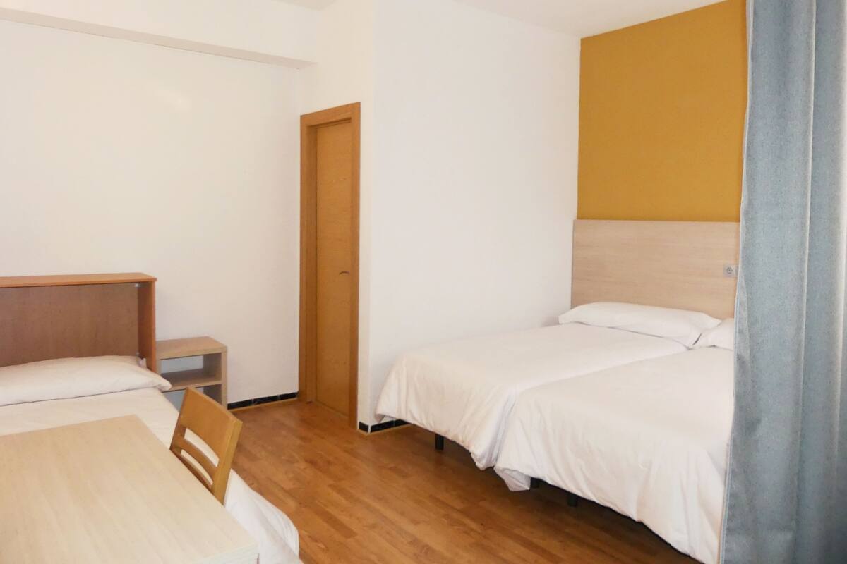 Una habitación pequeña y limpia con dos camas, una mesa de madera y una silla.