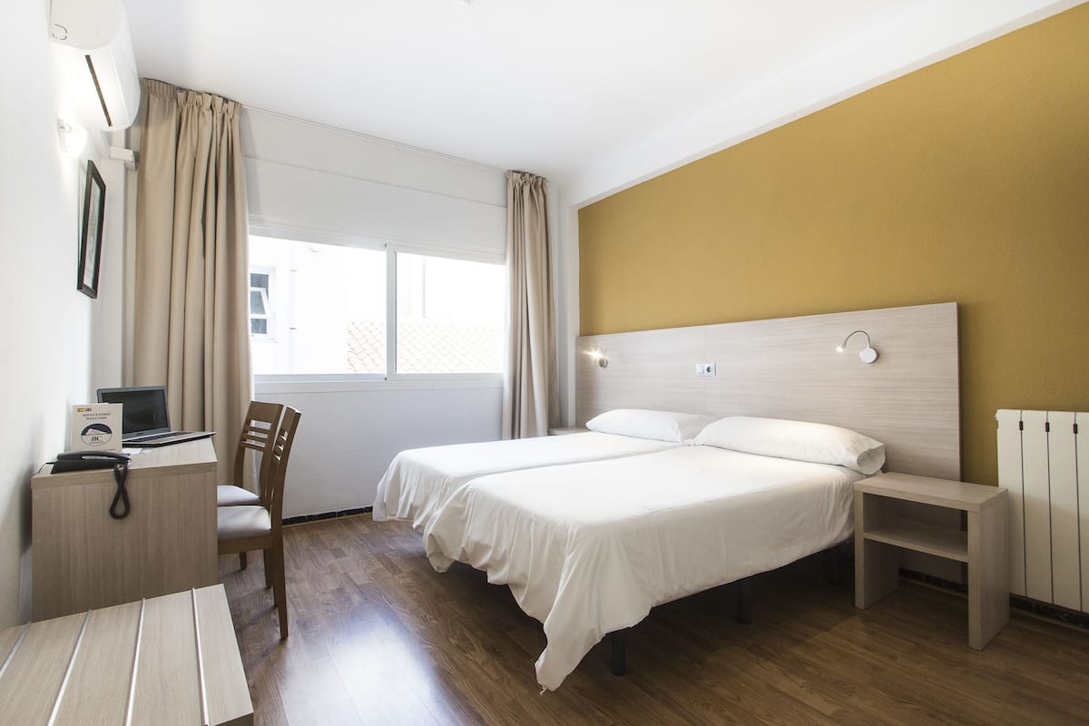 Una habitación de hotel con cama, escritorio, silla, mesita de noche y ventana con cortinas.