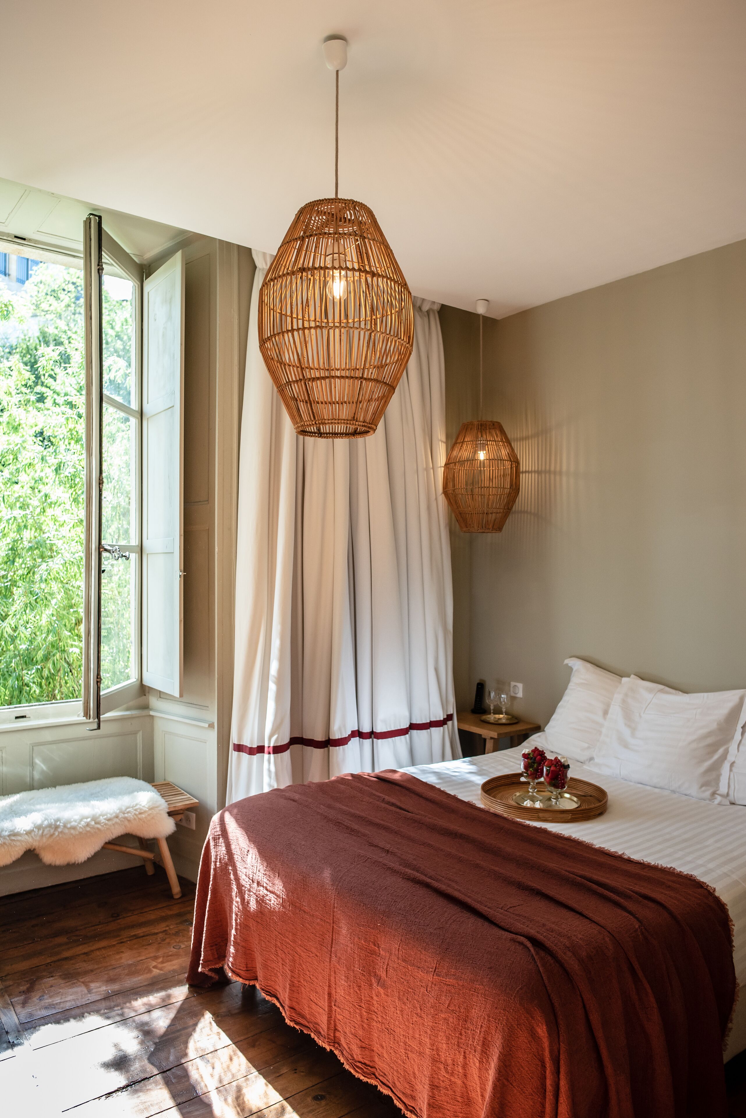 A bedroom with a large window, a wooden floor, a bed with a red blanket, and two wicker pendant lights.