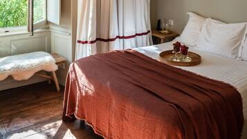 Un dormitorio con un ventanal grande, suelo de madera, una cama con una manta roja y dos lámparas colgantes de mimbre.