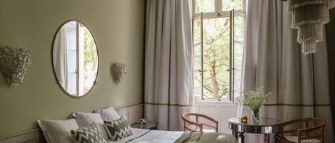 Un dormitorio con una cama, un espejo redondo, una ventana con cortinas, una mesa con un jarrón y dos sillas de mimbre.