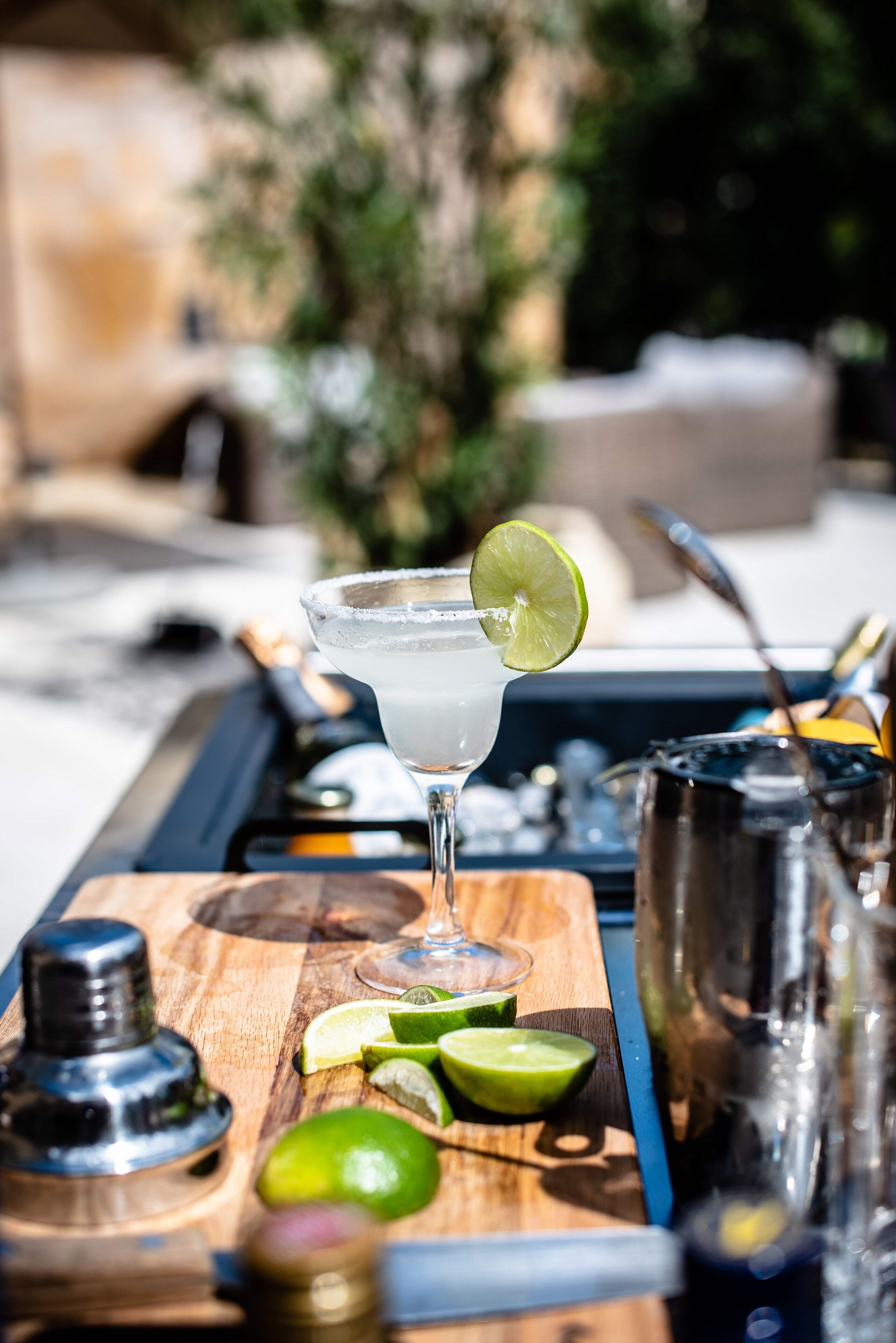 Een cocktailglas met een limoen, op een houten dienblad met een cocktailshaker, limoenpartjes en een fles.
