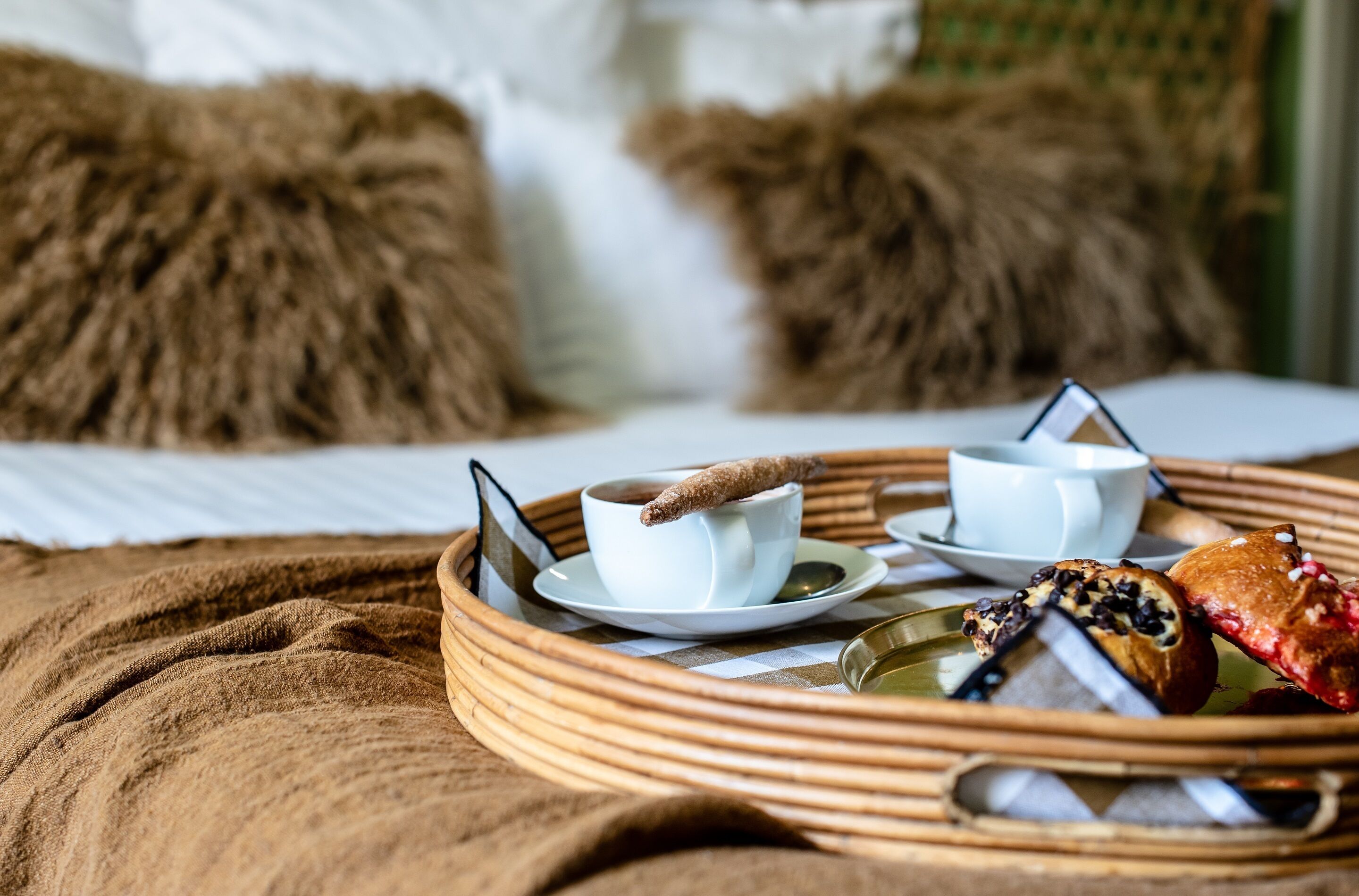 A breakfast tray with a cup of coffee, a pastry, and a teapot on a bed with a brown blanket and two pillows.