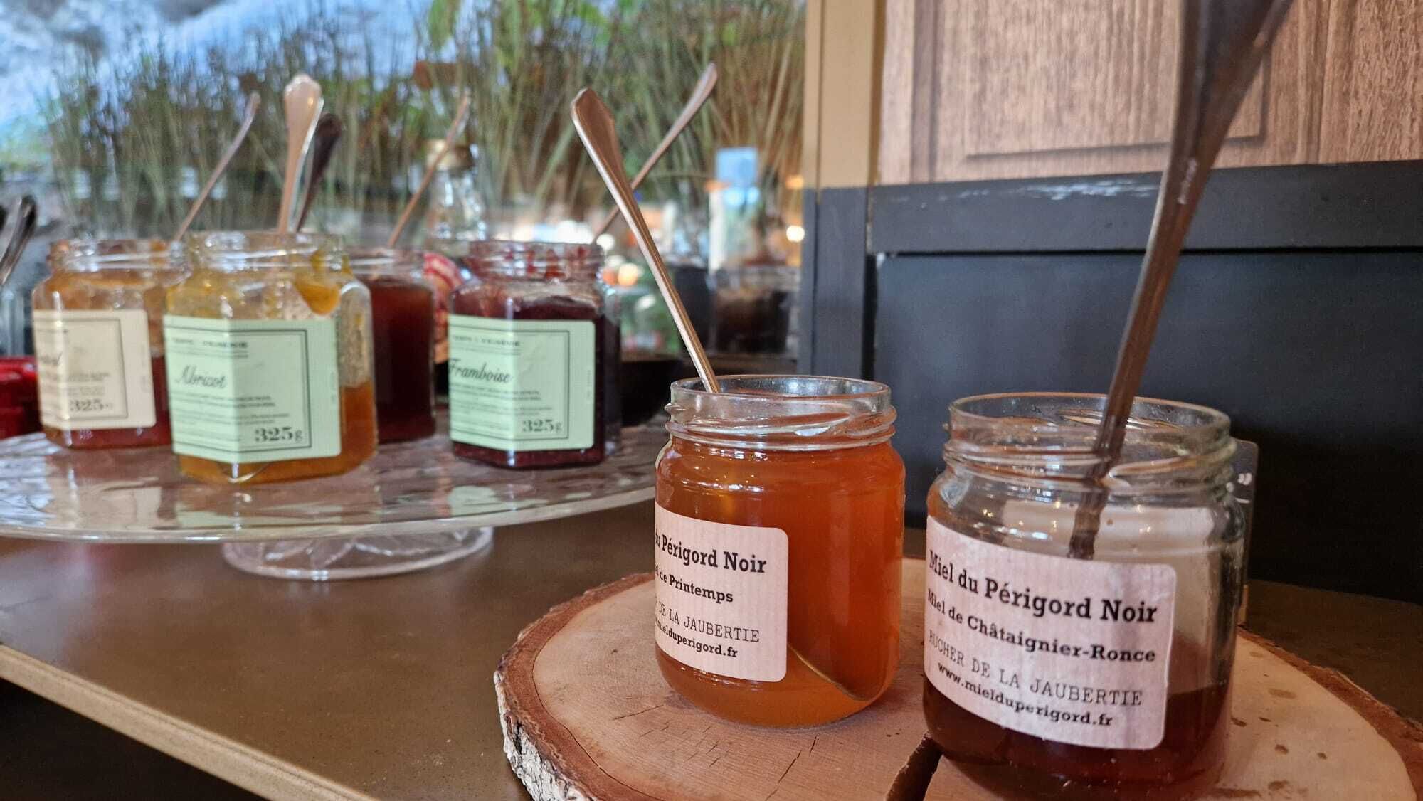 Jars of honey on a wooden board with labels, a spoon, and a window with a view of trees.