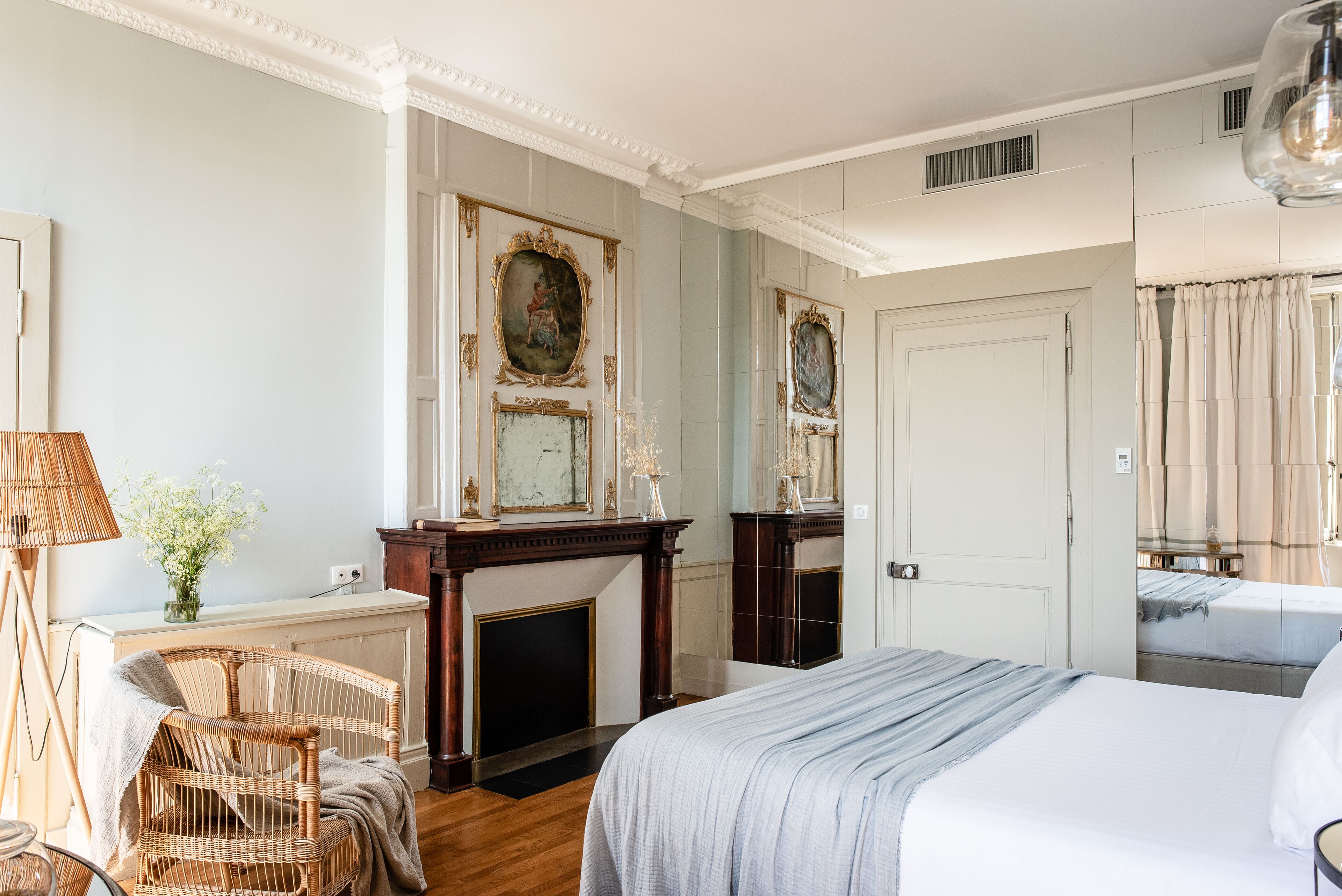 Une chambre d’hôtel avec un grand lit, un fauteuil en osier, une cheminée et un miroir.