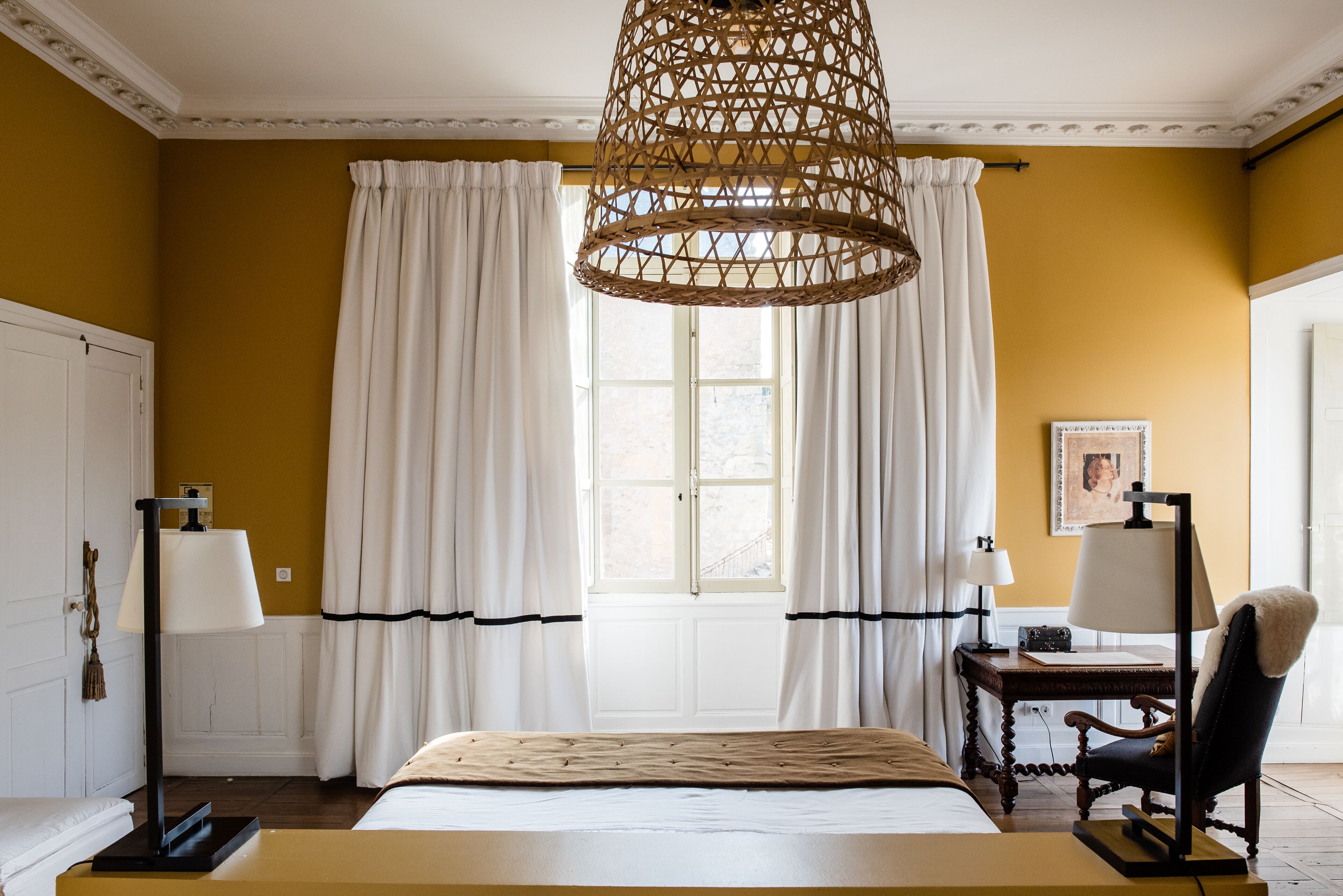 A bedroom with a bed, a desk with a lamp, a chair, and a window with curtains.