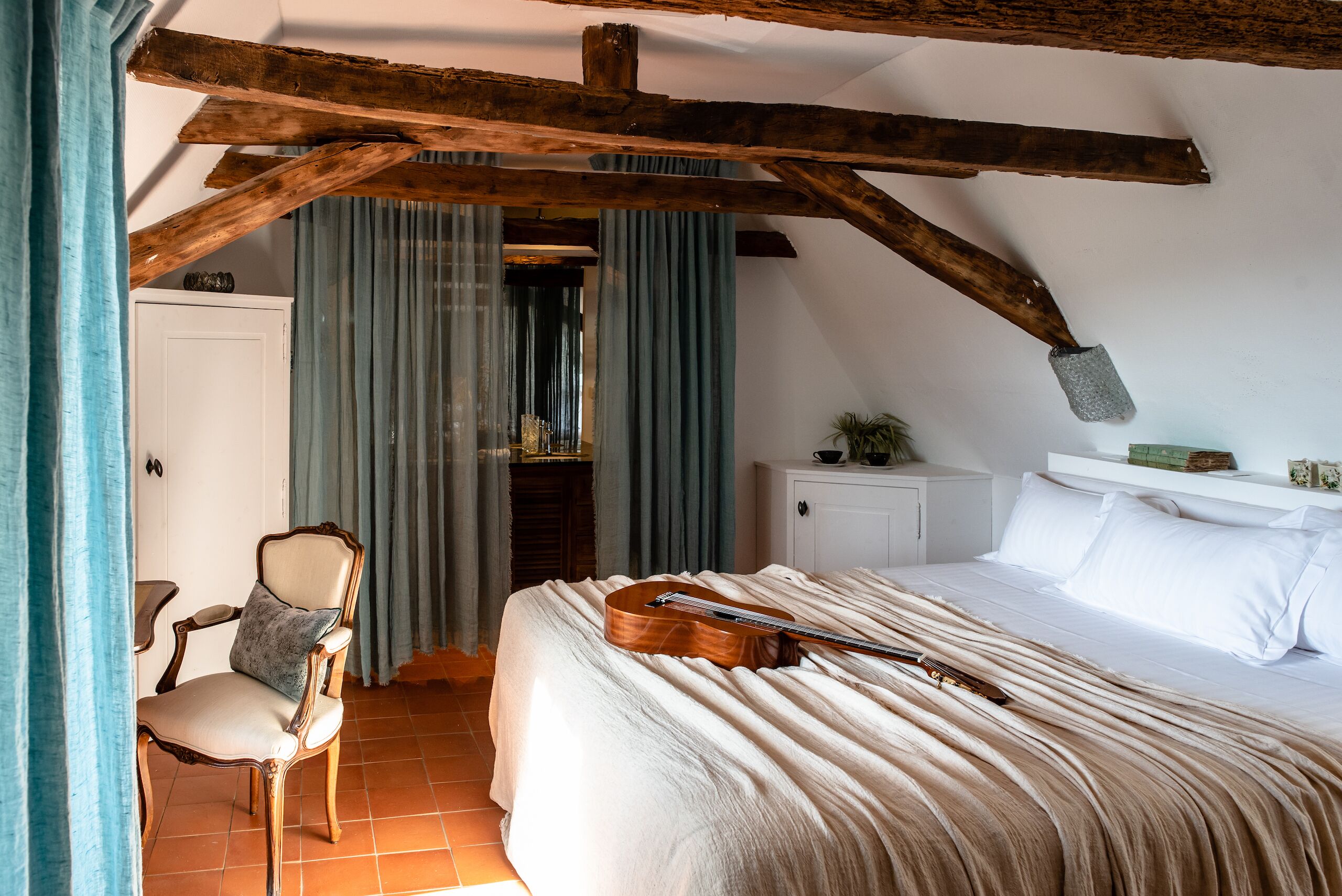 Quarto com teto de madeira, uma cama com um violão, uma cadeira e uma janela com cortinas.