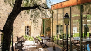 An outdoor patio with tables and chairs, a large tree, and a building with large windows. - Hôtel de Bouilhac (Montignac)