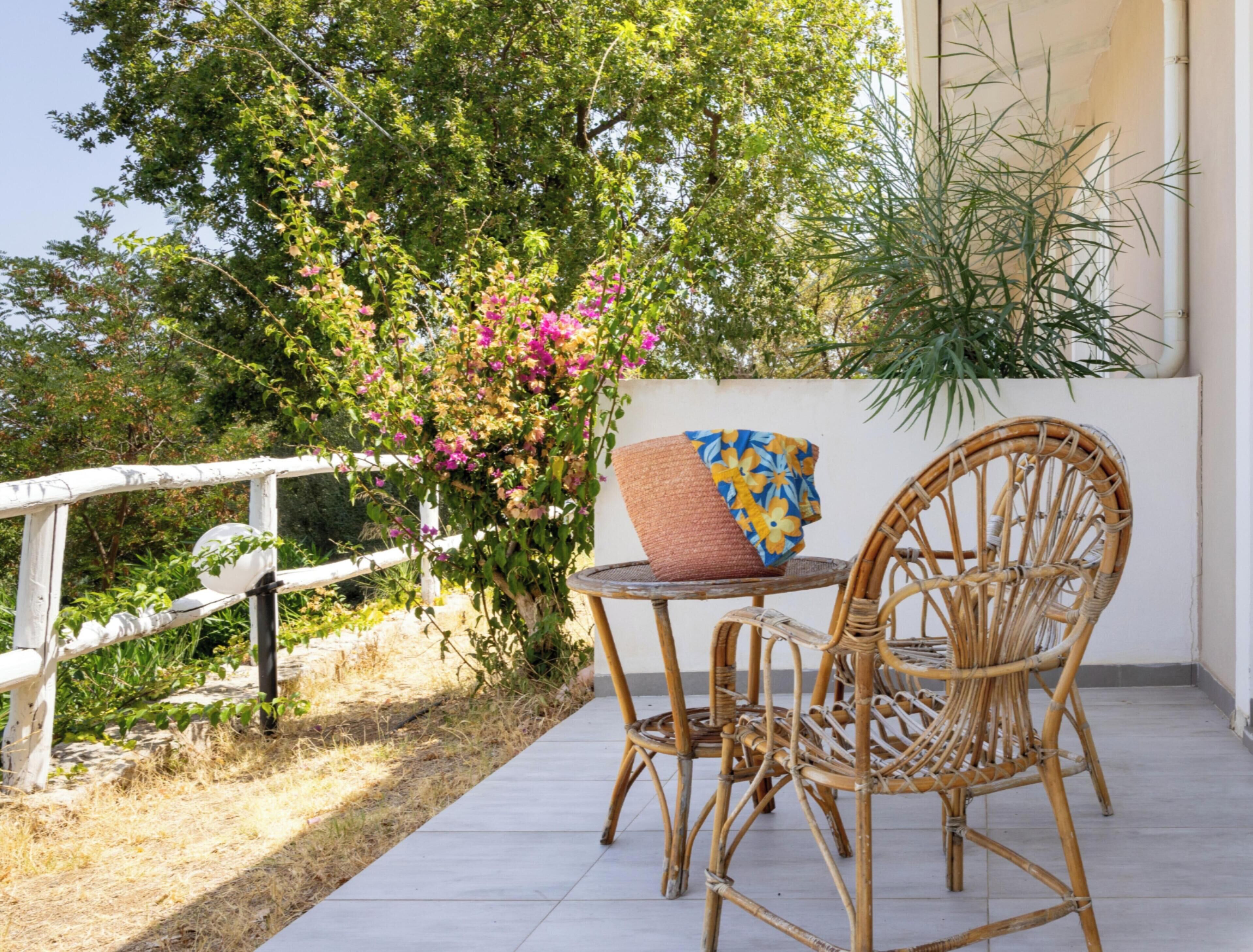 Una silla de mimbre y una mesa en un balcón con baranda blanca, una planta en maceta y un arbusto en flor.