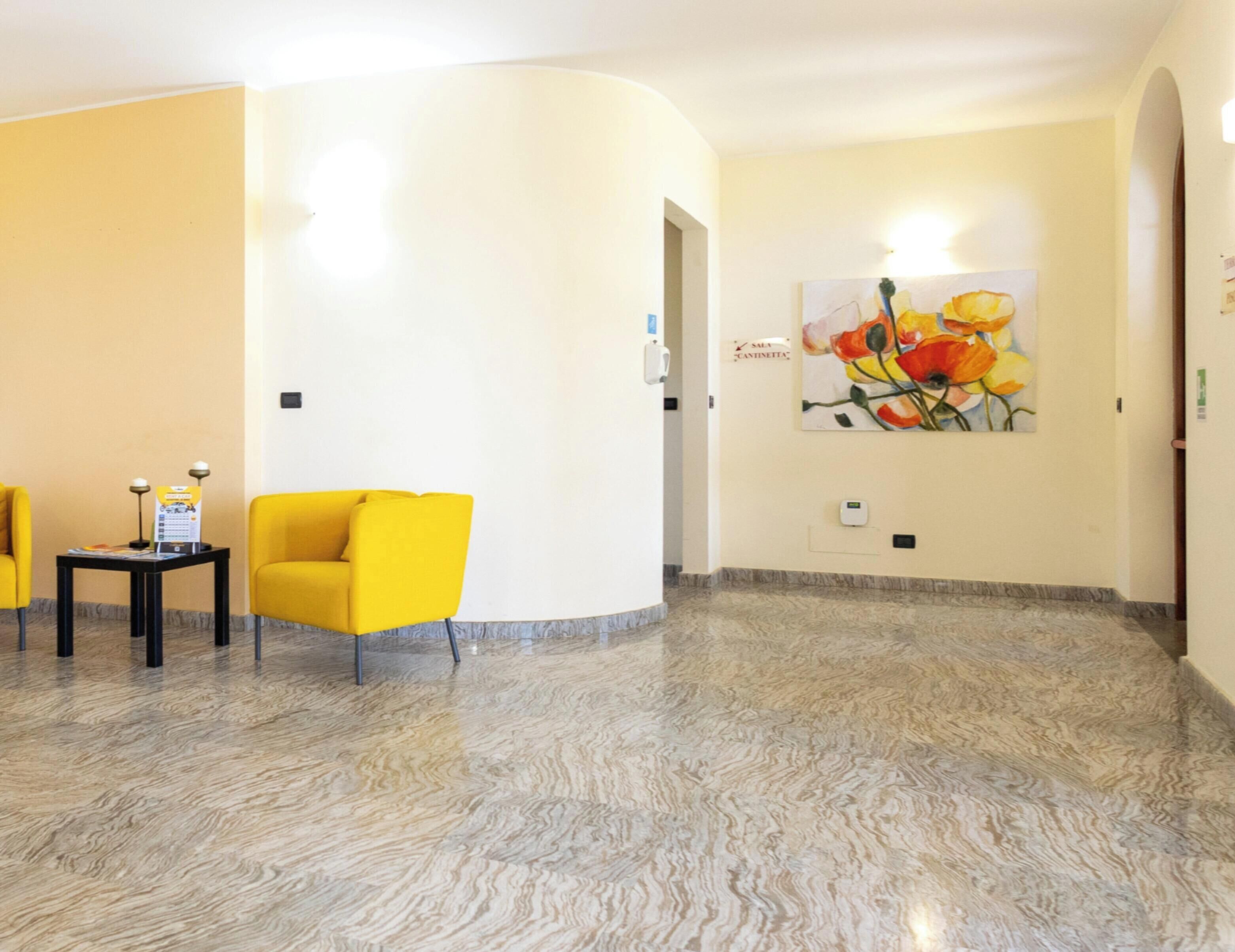 A hotel lobby with a marble floor, yellow chairs, a small table with a lamp, and a painting of flowers on the wall.
