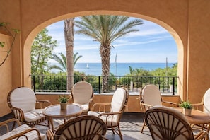 Ein Balkon mit Korbstühlen und einem Tisch, der Blick auf die Küste mit Palmen und dem Meer bietet.