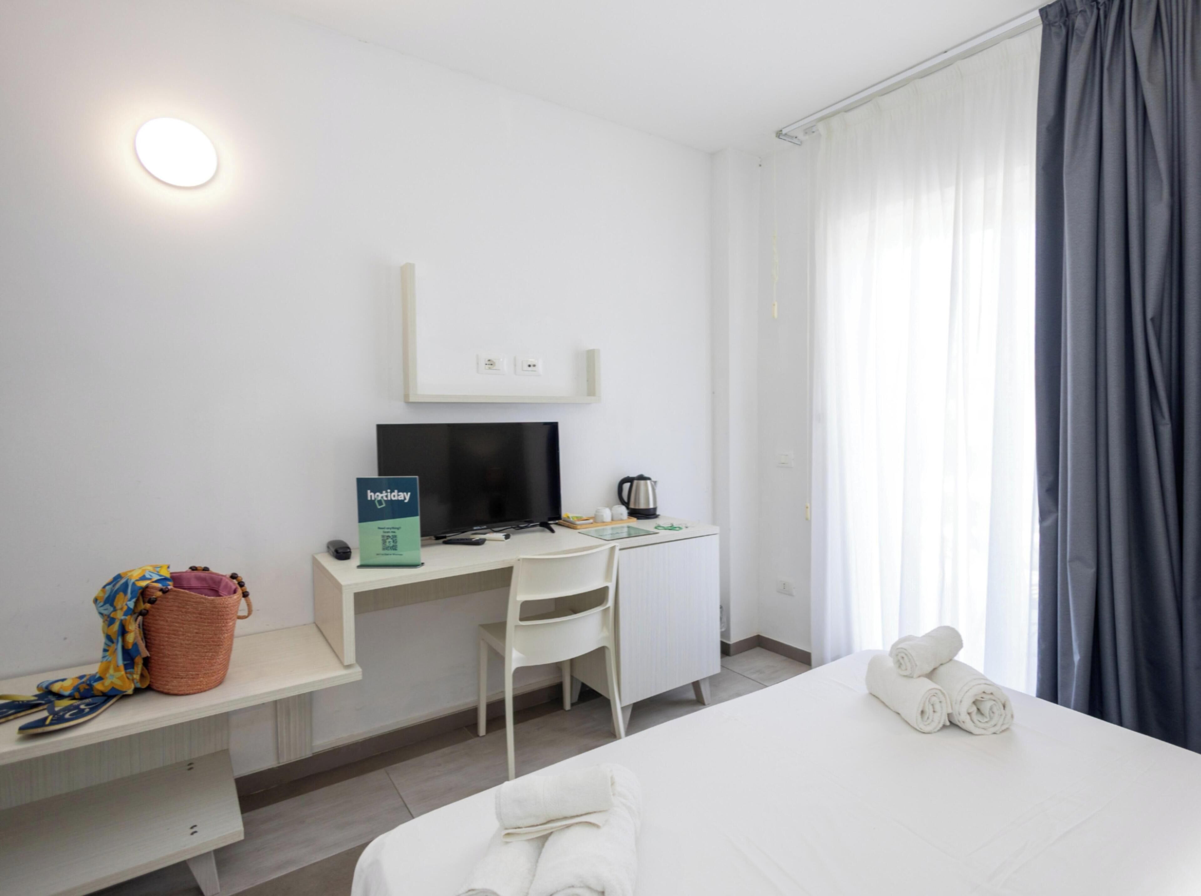Habitació d'hotel amb llit, escriptori amb ordinador, cadira, taula petita amb cistella i finestra amb cortines.