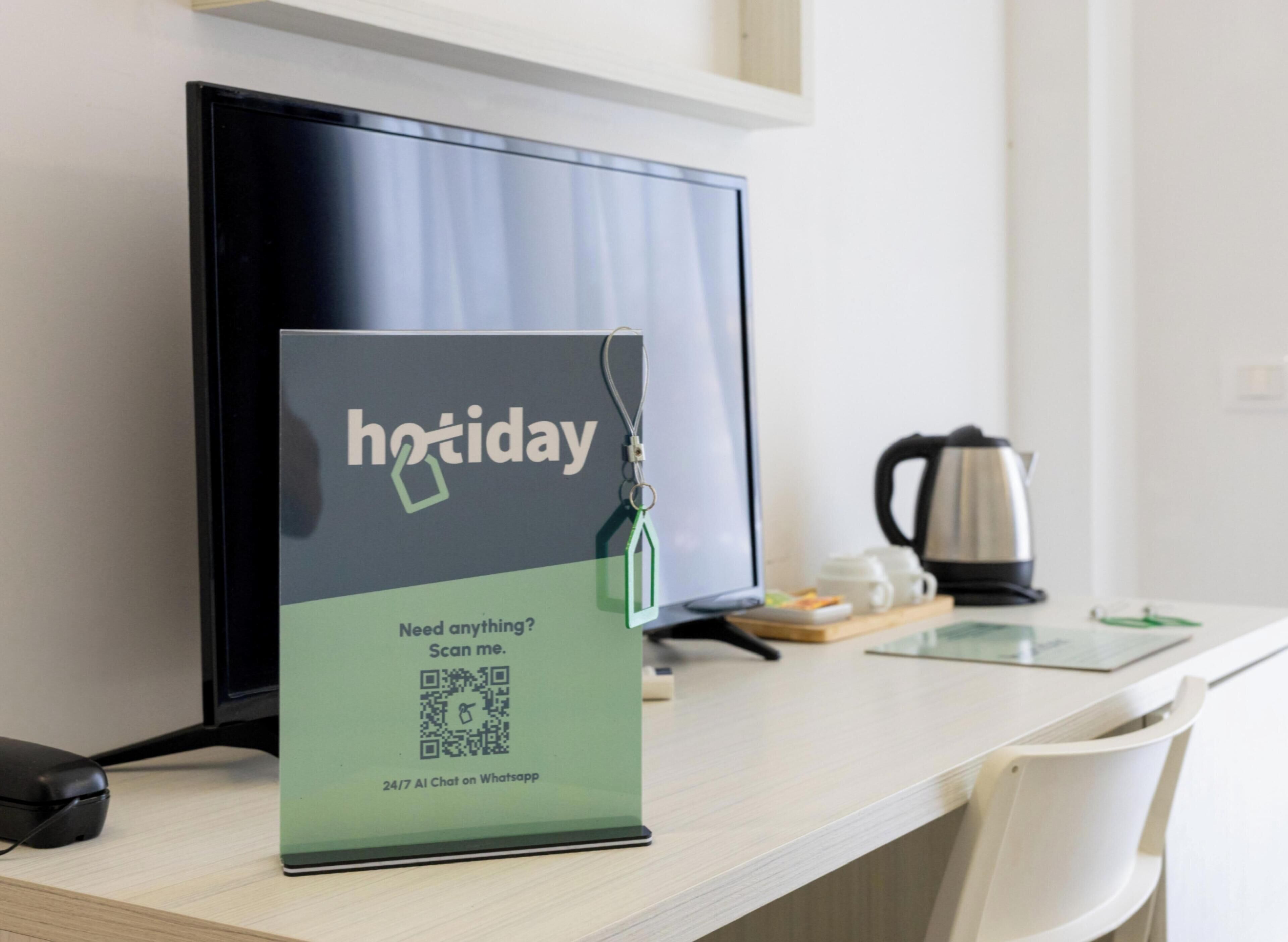 Mostrador de hotel con un monitor mostrando un letrero que dice 'vacaciones' y un código QR para un chat con IA las 24 horas, los 7 días de la semana en WhatsApp.