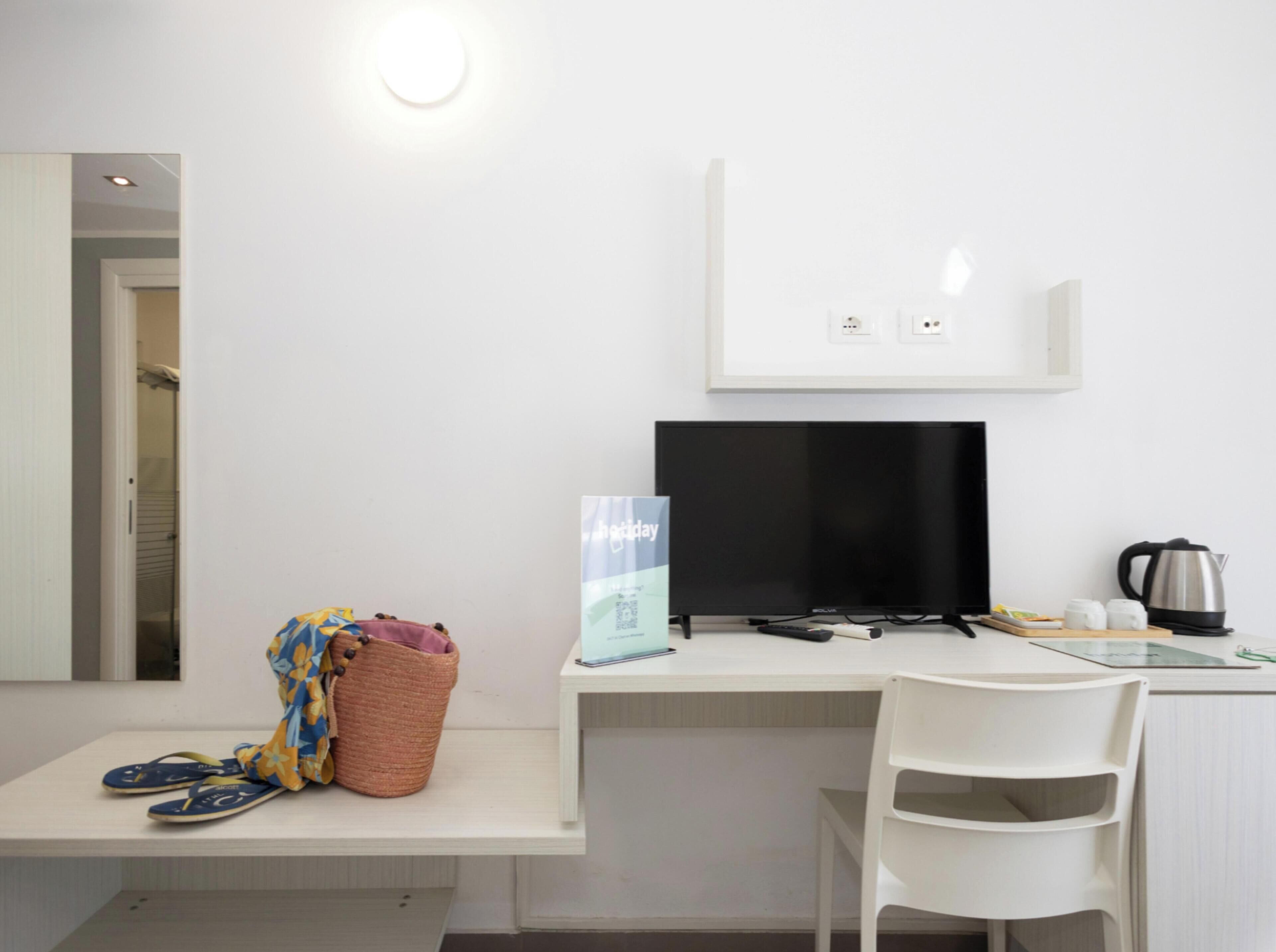 A room with a desk, a television, a basket, a pair of flip-flops, and a kettle.