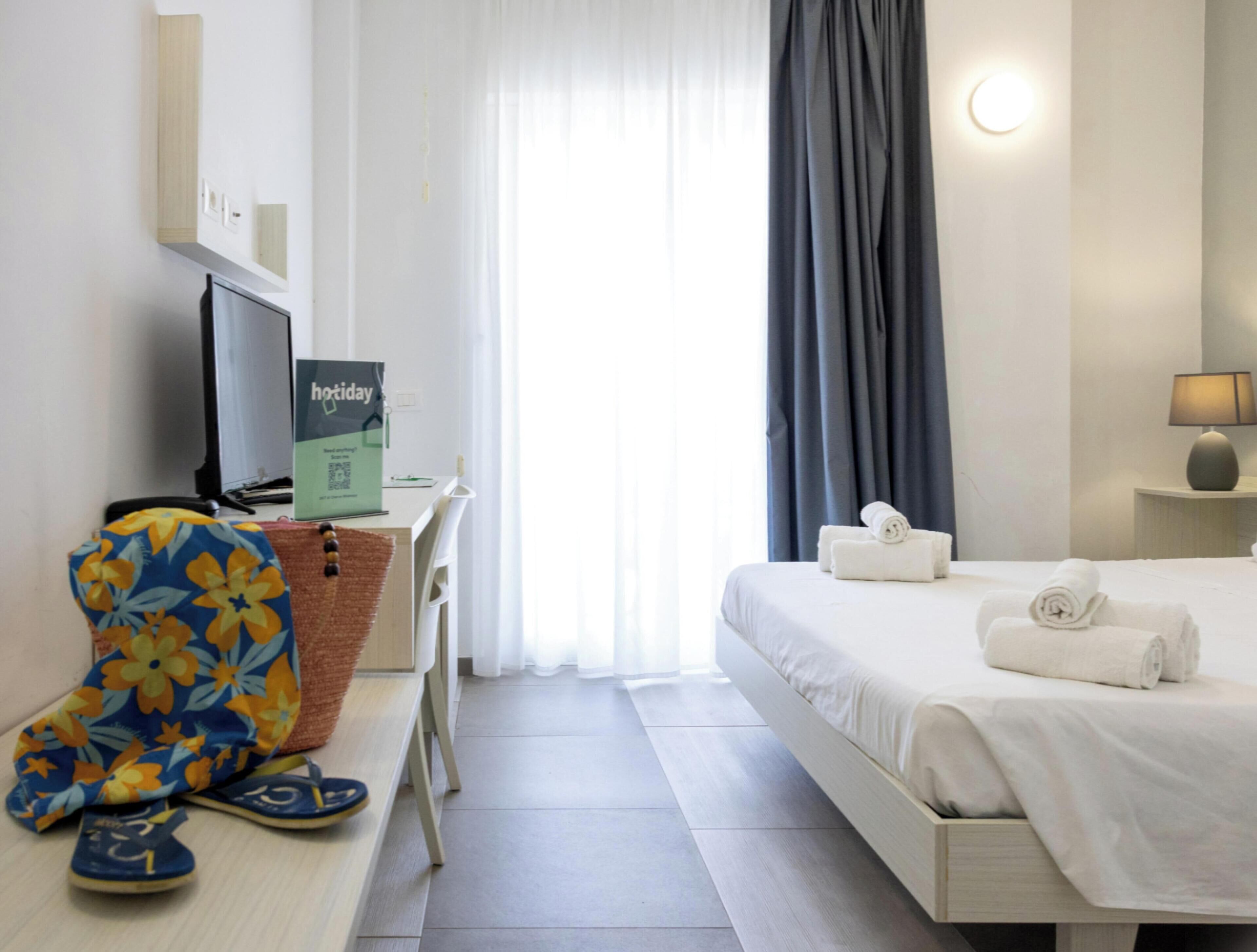 Una habitación de hotel con cama, escritorio, computadora, silla y ventana con cortinas.