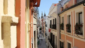 Una calle estrecha flanqueada por edificios, con balcones adornados con plantas y un cielo despejado.