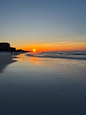 A beach with a sunset, calm water, and buildings in the background.