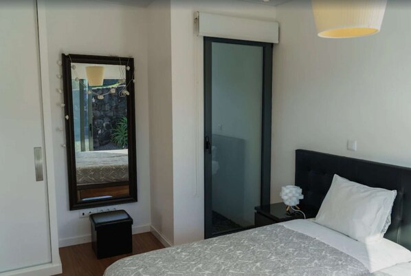 A bedroom with a bed, a bedside table, a mirror, and a door leading to a balcony. - Apartment with garden view (Angra do Heroísmo)