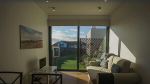 Una sala de estar moderna con un sofá, una mesita y un televisor. Hay un ventanal corredero que da a un jardín con cerca y una casa al fondo.