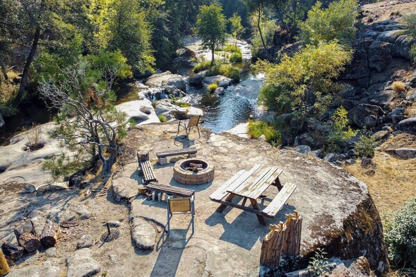 A rustic outdoor seating area with a fire pit, benches, and a table, surrounded by rocks and trees near a stream.