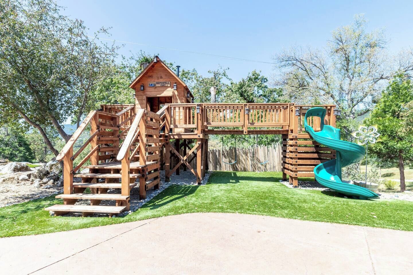A wooden playground structure with a slide and stairs, surrounded by trees and a clear sky.