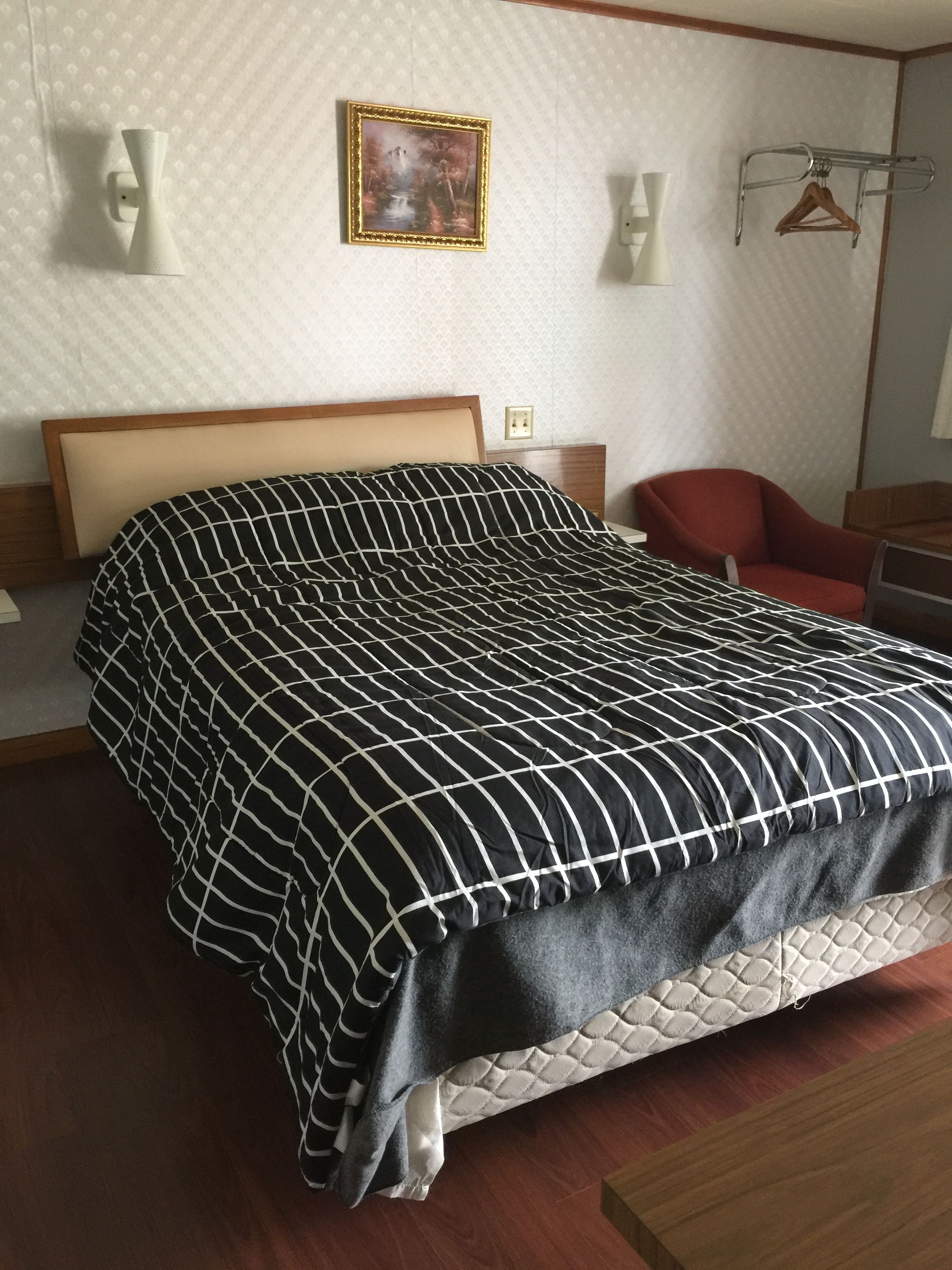 A neatly made bed with a black and white checkered bedspread, a wooden headboard, and a quilted mattress. There is a red chair and a wooden nightstand with a lamp.