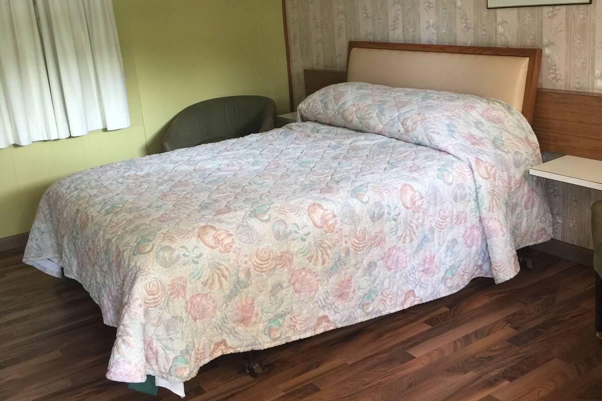 A hotel room with a wooden floor, a bed with floral bedding, a green chair, a bedside table, a lamp, and a floral painting on the wall.