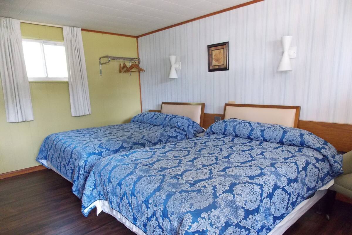 A double bed with a blue patterned bedspread, a wooden headboard, a wall-mounted lamp, and a window with curtains.