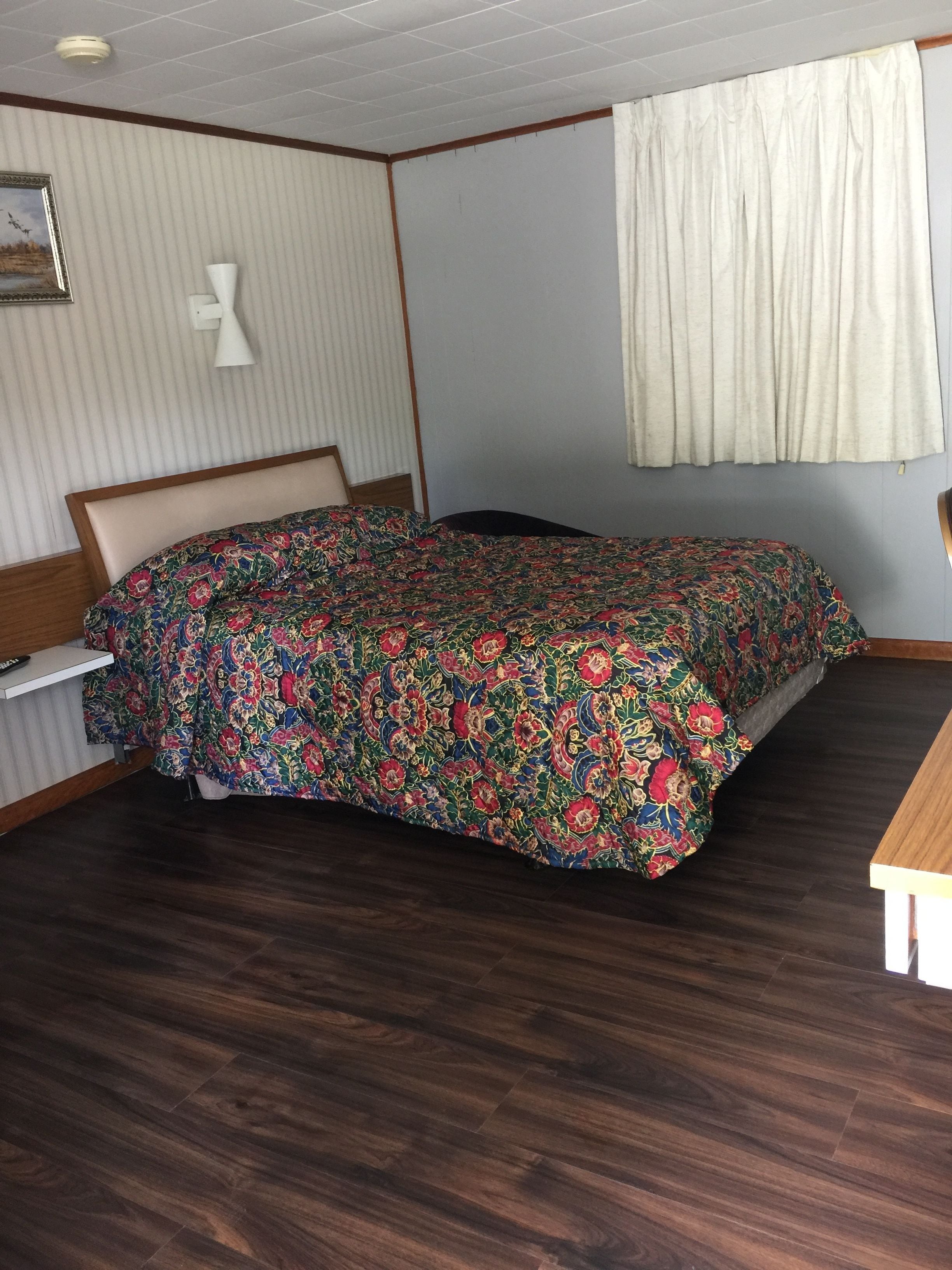 Un dormitorio con cama, mesita de noche, lámpara, ventana con cortinas e imagen colgada en la pared.