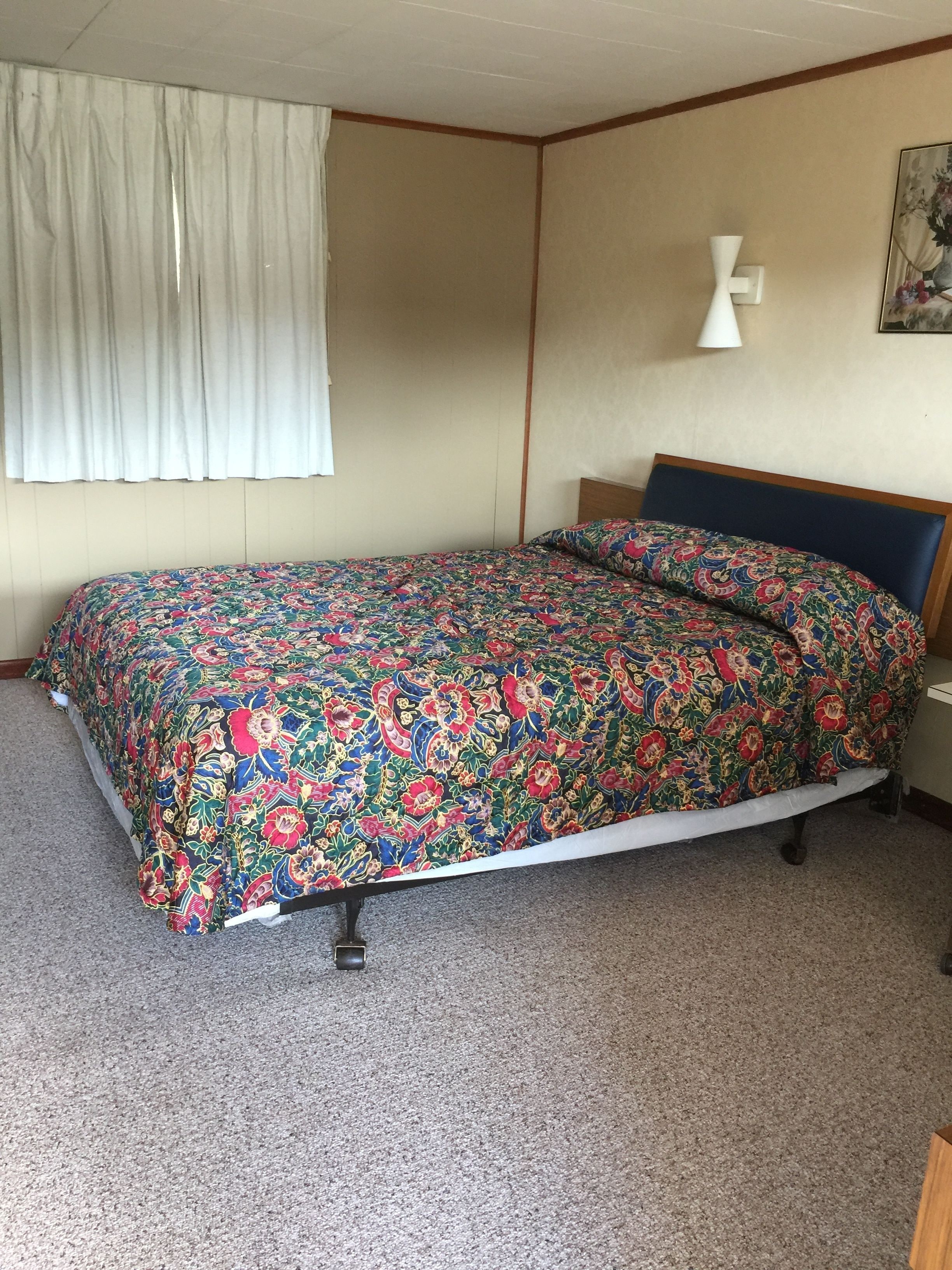 Et soveværelse med blomstret sengetæppe, sengegavl i træ, hvid lampe og et billede på væggen.