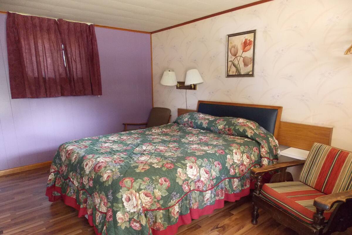 A bedroom with a floral bedspread, a wooden headboard, a wooden chair, a nightstand, and a wall-mounted lamp.