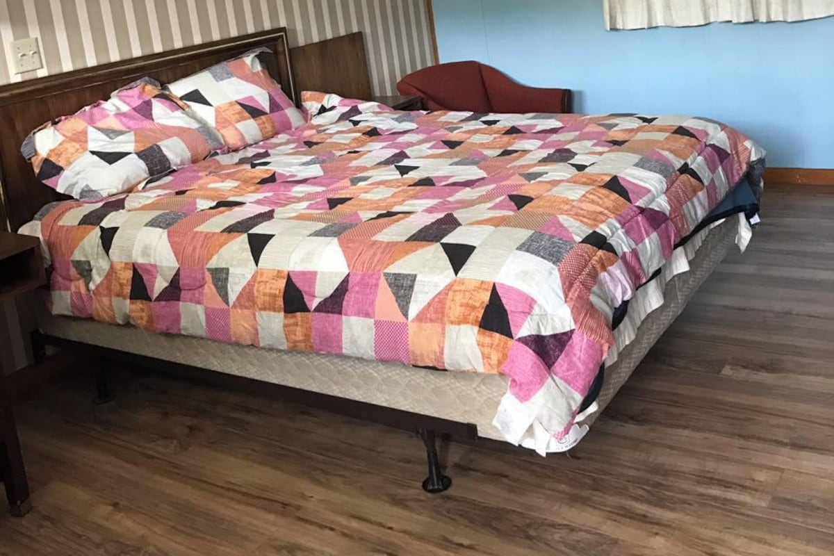 A bedroom with a large bed, wooden headboard, striped wallpaper, a painting, a ceiling light, and a window with curtains.