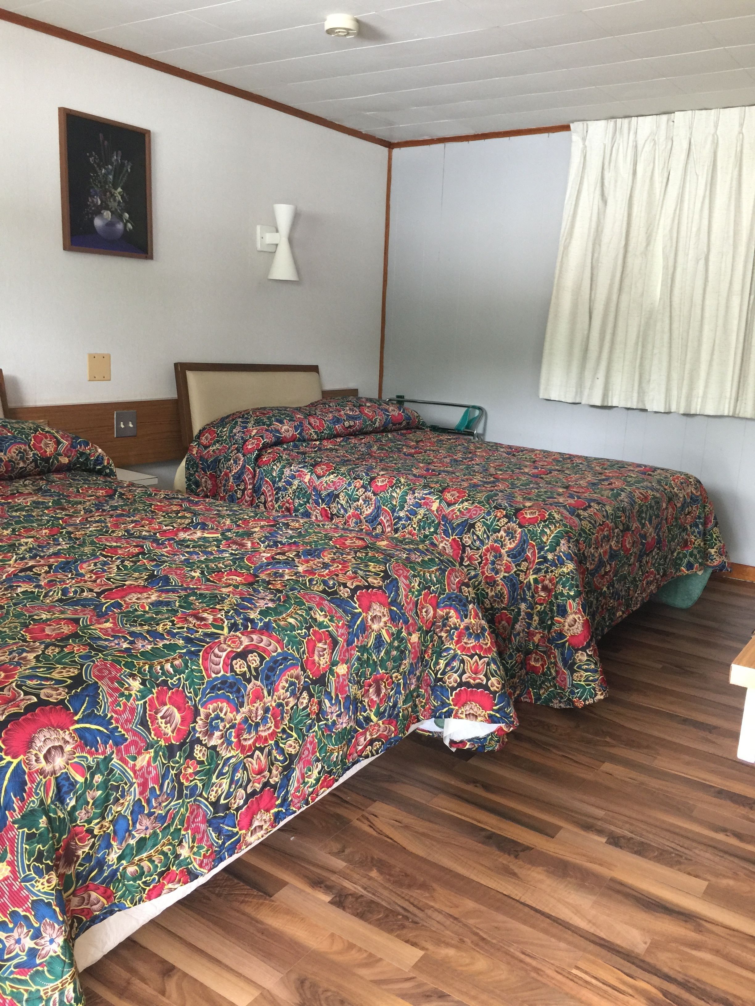 Un dormitorio con dos camas, suelo de madera, pared blanca, un cuadro enmarcado y una ventana con cortinas.