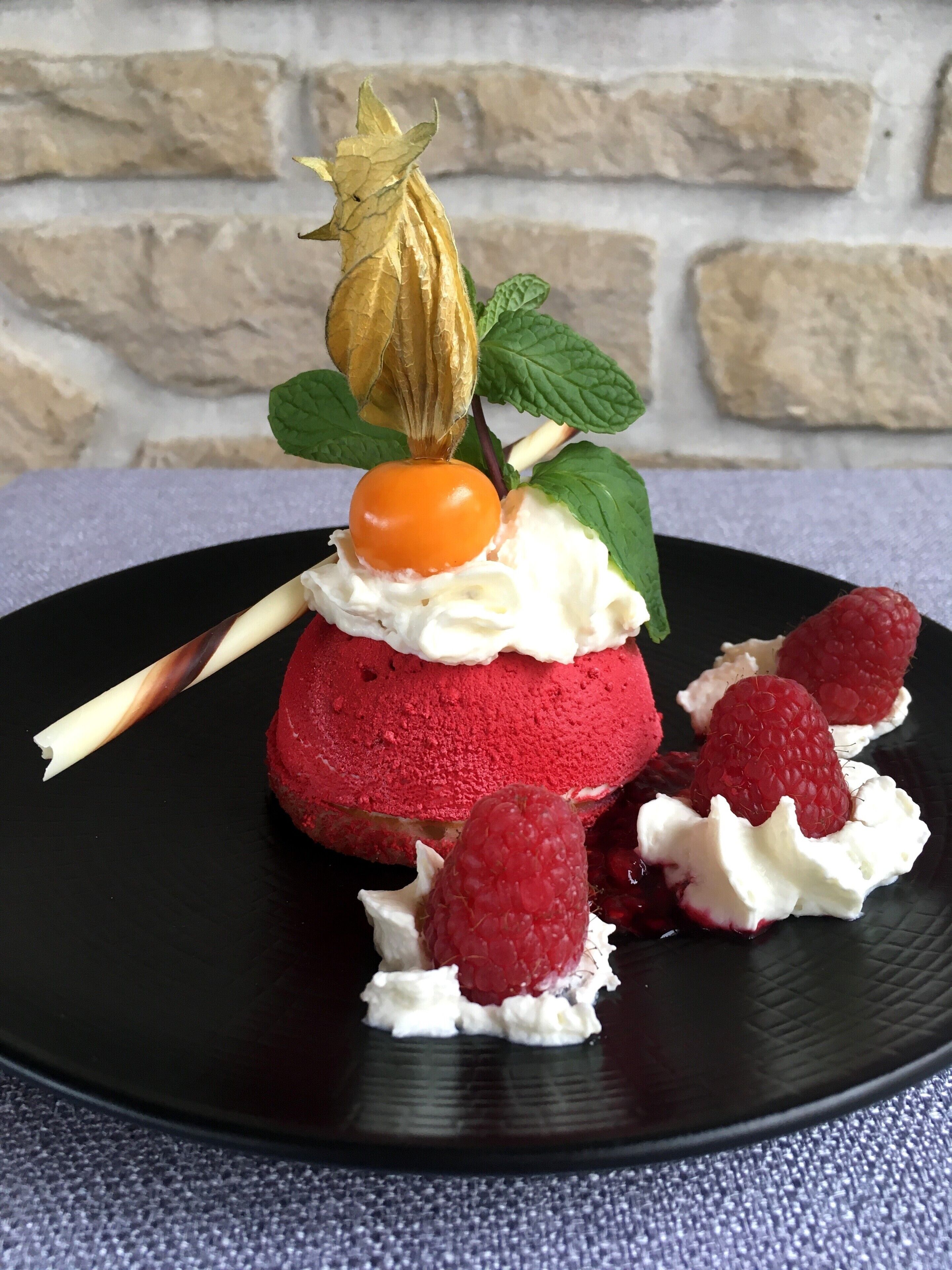 A dessert with a red base, topped with whipped cream and a cherry, garnished with mint leaves and a dried flower.