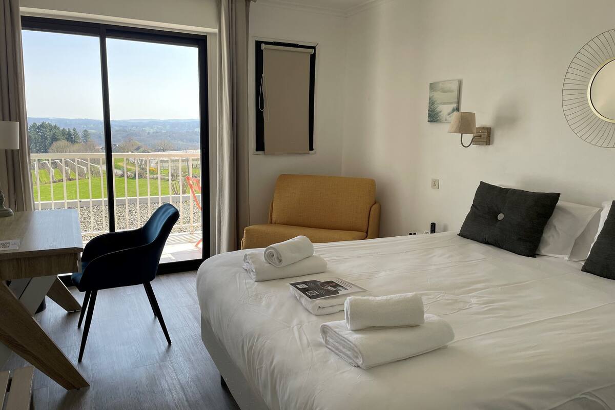 A hotel room with a large bed, a desk, a chair, and a view of a green landscape through a large window.