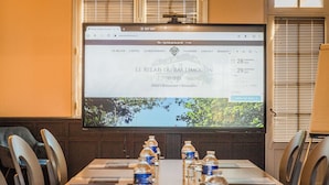 Habitación de hotel con una pantalla grande mostrando un sitio web, una mesa de conferencias con botellas de agua y vistas a árboles por la ventana.