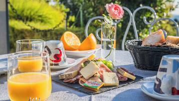 En frukostuppläggning med ost, charkuterier och frukt på ett bord, med en ros i en vas och en korg med bröd.