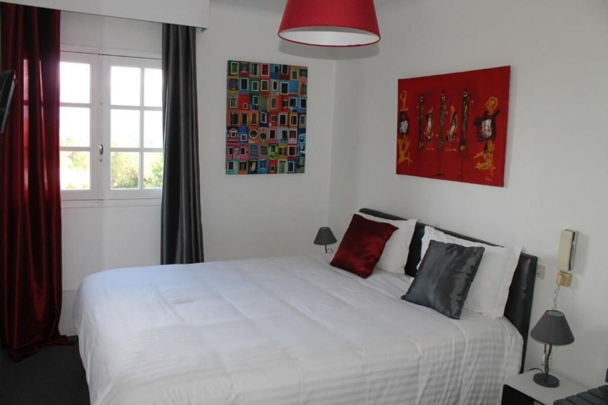 Un dormitorio con cama, ventana con cortinas, una lámpara roja y dos obras de arte en las paredes.